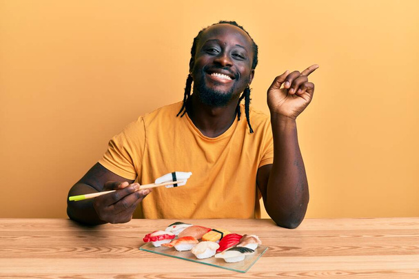 Knappe jonge zwarte man eet sushi zittend op de tafel lachend vrolijk wijzend met hand en vinger naar de zijkant  - Foto, afbeelding