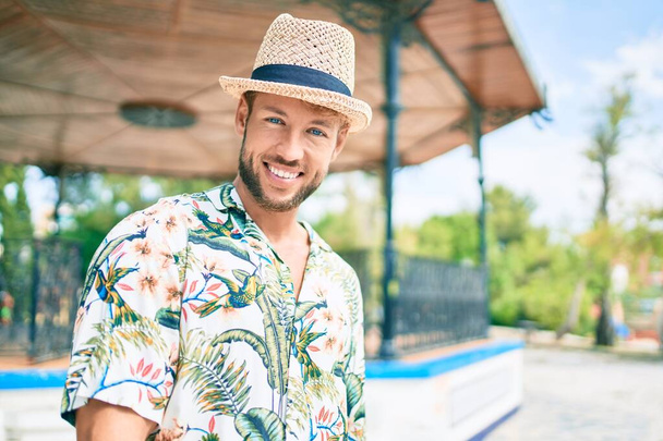 Handsome caucasian man wearing summer hat and flowers shirt smiling happy outdoors - 写真・画像