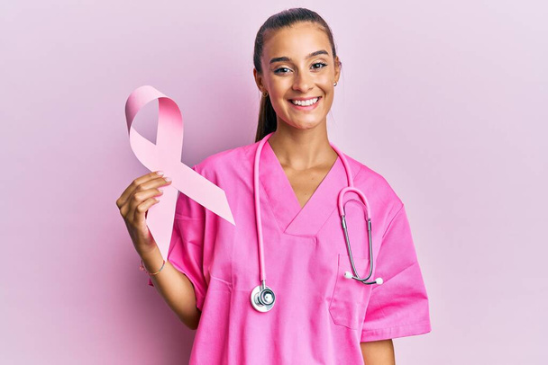 Jeune femme hispanique tenant un ruban de cancer rose regardant positif et heureux debout et souriant avec un sourire confiant montrant des dents  - Photo, image