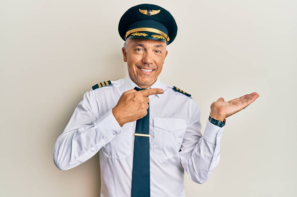 Knappe middelbare leeftijd volwassen man dragen vliegtuig piloot uniform verbaasd en glimlachen naar de camera terwijl presenteren met de hand en wijzen met de vinger.  - Foto, afbeelding