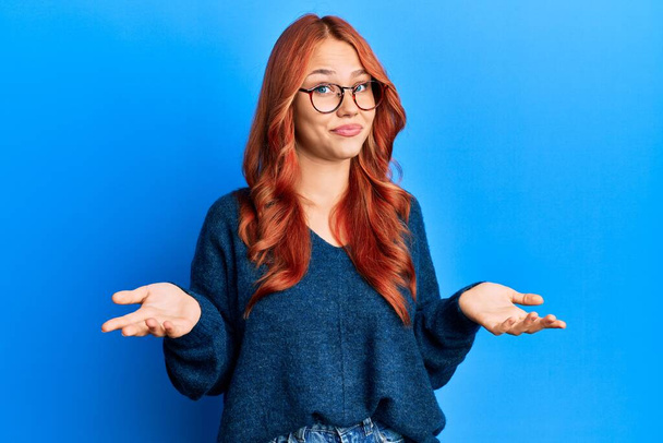 Junge schöne rothaarige Frau in lässigem Pullover und Brille vor blauem Hintergrund ahnungslos und verwirrt mit erhobenen Armen und Händen. Zweifelhaftes Konzept.  - Foto, Bild