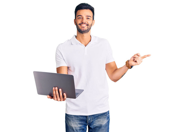 Joven hombre hispano guapo trabajando usando computadora portátil sonriendo feliz señalando con la mano y el dedo a un lado  - Foto, Imagen