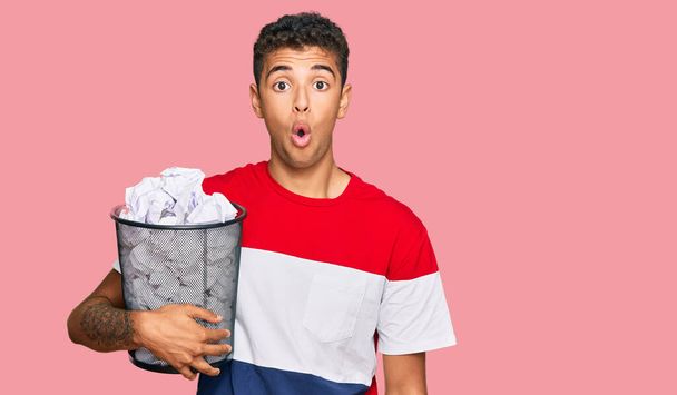 Young handsome african american man holding paper bin full of crumpled papers scared and amazed with open mouth for surprise, disbelief face  - Photo, Image