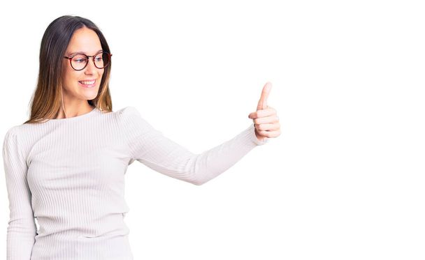 Beautiful brunette young woman wearing casual white sweater and glasses looking proud, smiling doing thumbs up gesture to the side  - Photo, Image