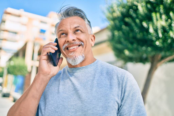 Middelbare leeftijd hispanic grijs-harige man glimlachend gelukkig praten op de smartphone in de stad. - Foto, afbeelding