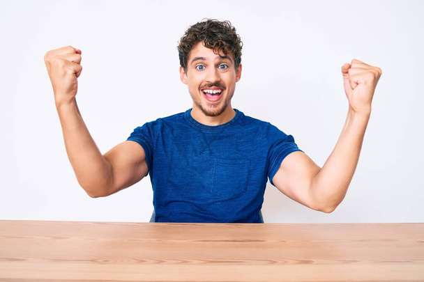 Young caucasian man with curly hair wearing casual clothes sitting on the table screaming proud, celebrating victory and success very excited with raised arms  - Foto, Bild