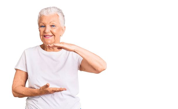 Senior beautiful woman with blue eyes and grey hair wearing casual white tshirt gesturing with hands showing big and large size sign, measure symbol. smiling looking at the camera. measuring concept.  - Foto, Imagen