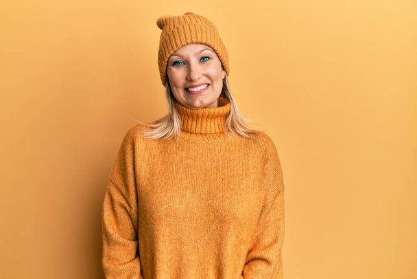 Mulher caucasiana de meia idade usando suéter de inverno de lã e chapéu olhando positivo e feliz de pé e sorrindo com um sorriso confiante mostrando dentes  - Foto, Imagem