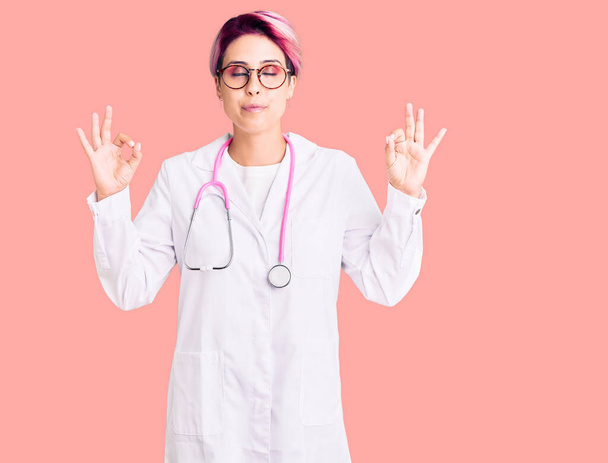 Молодая красивая женщина с розовыми волосами в докторской форме расслабляется и улыбается с закрытыми глазами, делая медитационный жест пальцами. Концепция йоги.  - Фото, изображение