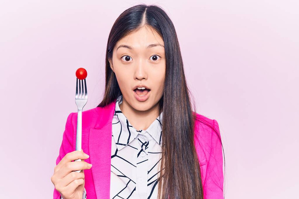Young beautiful chinese woman holding fork with tomato scared and amazed with open mouth for surprise, disbelief face  - Photo, Image