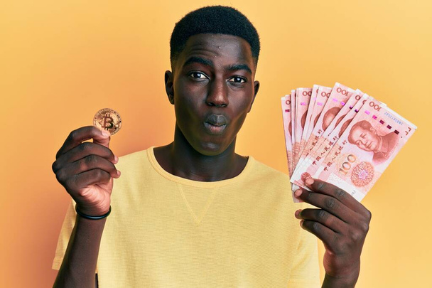 Young african american man holding yuan chinese banknotes and bitcoin making fish face with mouth and squinting eyes, crazy and comical.  - Фото, зображення