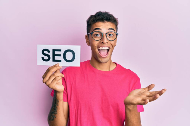 Young handsome african american man holding seo message paper celebrating achievement with happy smile and winner expression with raised hand  - Foto, Imagem