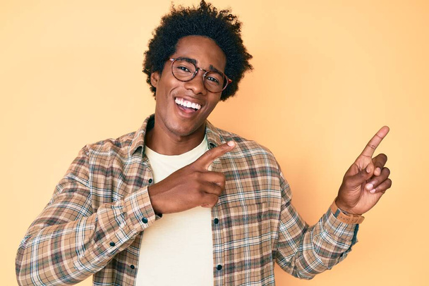 Hombre afroamericano guapo con pelo afro vistiendo ropa casual y gafas sonriendo y mirando a la cámara apuntando con dos manos y dedos hacia un lado.  - Foto, Imagen