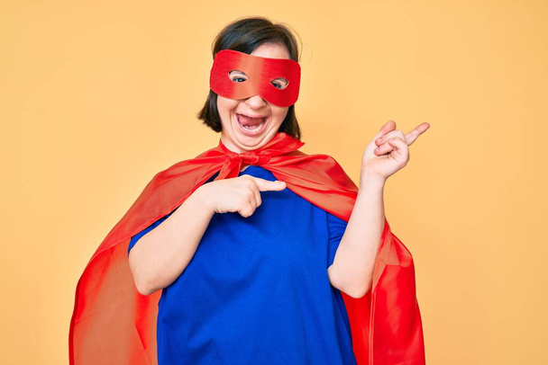 Mujer morena con síndrome de Down vistiendo disfraz de superhéroe sonriendo y mirando a la cámara apuntando con dos manos y dedos hacia un lado.  - Foto, Imagen