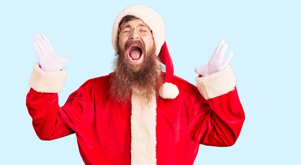 Handsome young red head man with long beard wearing santa claus costume celebrating mad and crazy for success with arms raised and closed eyes screaming excited. winner concept  - Valokuva, kuva