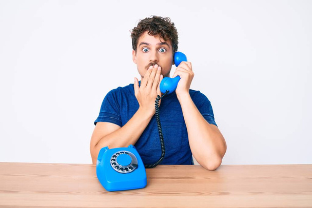 Young caucasian man with curly hair using vintage telephone sitting on the table covering mouth with hand, shocked and afraid for mistake. surprised expression  - Foto, Imagem
