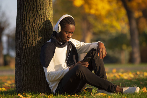 African man listening to music in headphones leaning on a tree trunk at the fall day - Foto, Imagen