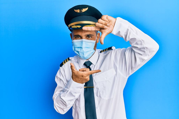 Handsome man with beard wearing airplane pilot uniform wearing safety mask smiling making frame with hands and fingers with happy face. creativity and photography concept.  - Foto, imagen