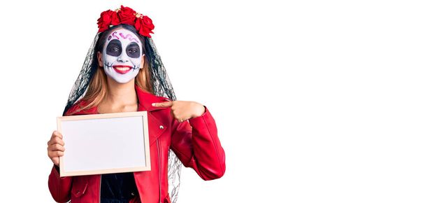 Woman wearing day of the dead costume holding empty white chalkboard pointing finger to one self smiling happy and proud  - Photo, Image