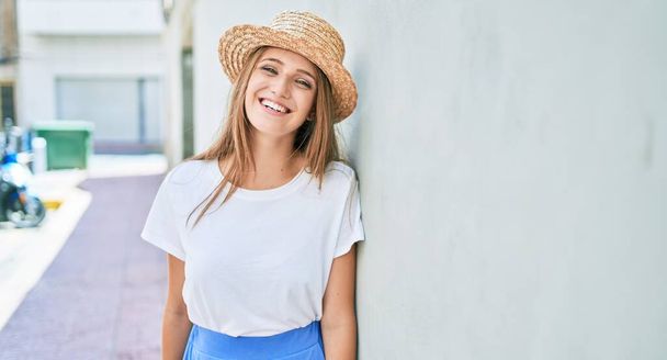 jonge blonde vrouw op vakantie glimlachen gelukkig leunend op de muur op straat van de stad - Foto, afbeelding