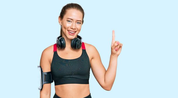 Beautiful blonde woman wearing gym clothes and using headphones pointing finger up with successful idea. exited and happy. number one.  - Fotó, kép