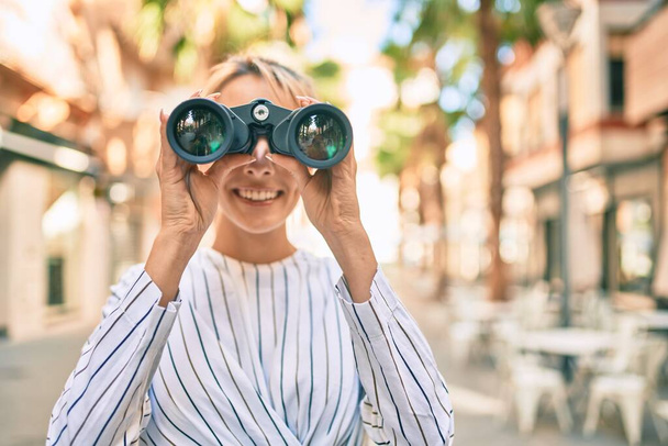 Joven mujer de negocios rubia sonriendo feliz usando prismáticos en la ciudad. - Foto, imagen