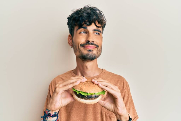 Giovane uomo ispanico mangiare un gustoso hamburger classico sorridente guardando di lato e fissando via pensando.  - Foto, immagini