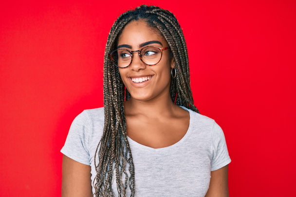 Νεαρή Αφροαμερικανή γυναίκα με κοτσίδες που φοράει casual ρούχα και γυαλιά κοιτάζοντας προς τα πλάγια, χαλαρώστε προφίλ ποζάρουν με φυσικό πρόσωπο και αυτοπεποίθηση χαμόγελο.  - Φωτογραφία, εικόνα