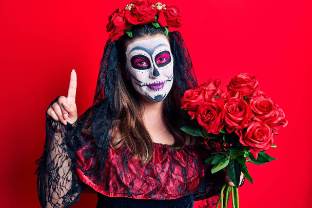 Jeune femme portant le jour du costume mort tenant des fleurs surpris par une idée ou une question pointant du doigt avec un visage heureux, numéro un  - Photo, image