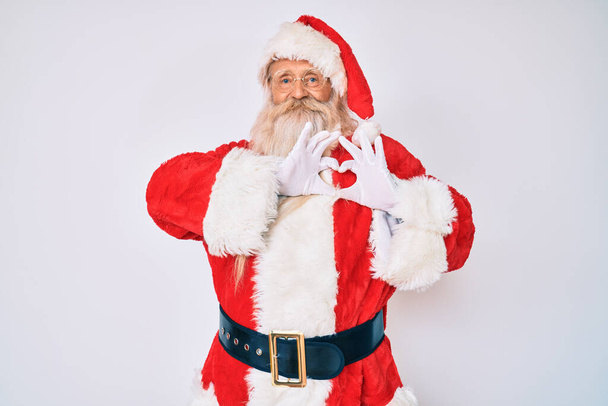 Vieil homme âgé aux cheveux gris et à la barbe longue portant le costume de Père Noël avec des bretelles souriant en amour faisant forme de symbole de coeur avec les mains. concept romantique.  - Photo, image