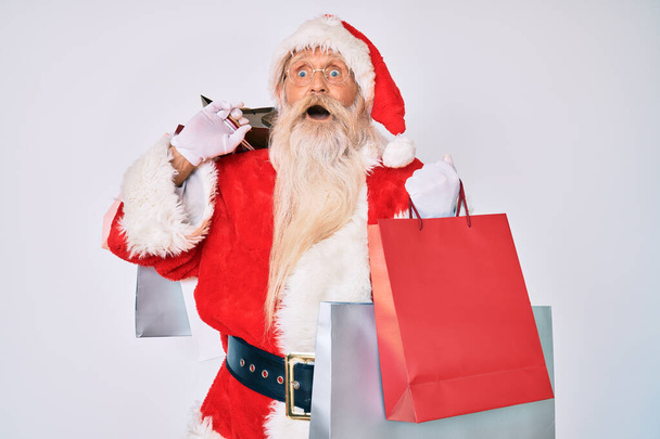 Gri saçlı ve uzun sakallı yaşlı bir adam Noel Baba kostümü giyiyor. Elinde alışveriş torbaları var. Çılgınca kutlama yapıyor..  - Fotoğraf, Görsel