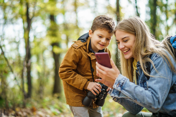 Madre e hijo felices están de excursión en el bosque. Están utilizando mapas en el teléfono inteligente. - Foto, Imagen