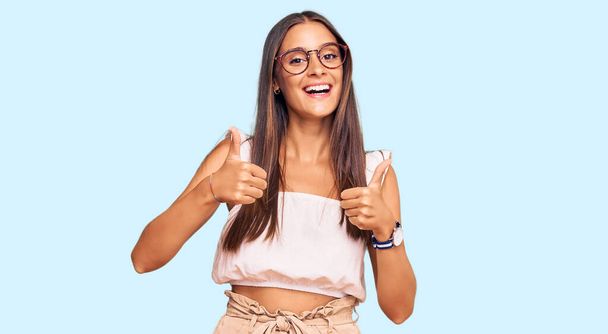 カジュアルな服やメガネの成功サインを身に着けている若いヒスパニック系の女性は手で積極的なジェスチャーを行う,親指を笑顔と幸せ.陽気な表情と勝者のジェスチャー.  - 写真・画像