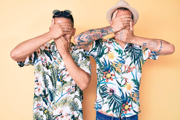 Fiatal meleg pár két férfi nyári kalapban és hawaii ingben, kezükkel befedik a szemüket és a szájukat, meglepve és sokkolva. rejtett érzelem  - Fotó, kép