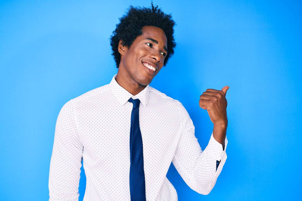 Schöner afrikanisch-amerikanischer Mann mit Afro-Haaren, Business-Hemd und Krawatte, Daumen zur Seite gerichtet, glücklich lächelnd mit offenem Mund  - Foto, Bild