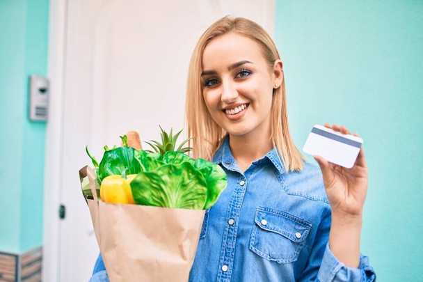 Młoda blondynka uśmiechnięta szczęśliwa trzymając torbę z zakupami i kartę kredytową stojącą w mieście. - Zdjęcie, obraz