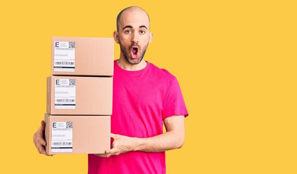 Young handsome man holding delivery package scared and amazed with open mouth for surprise, disbelief face  - Photo, Image