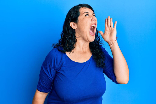 Além disso, mulher morena tamanho vestindo camisa azul casual gritando e gritando alto para o lado com a mão na boca. conceito de comunicação.  - Foto, Imagem