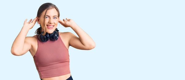 Piękna biała młoda kobieta w stroju siłowni i przy użyciu słuchawek uśmiechających się ciągnących uszy palcami, zabawny gest. problem przesłuchań  - Zdjęcie, obraz
