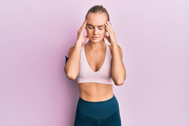 Beautiful blonde woman wearing sportswear and arm band with hand on head for pain in head because stress. suffering migraine.  - Foto, immagini