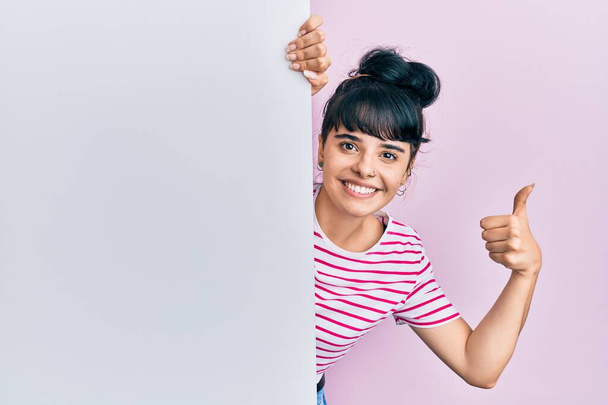 Menina hispânica jovem segurando banner vazio em branco sorrindo feliz e positivo, polegar para cima fazendo excelente e sinal de aprovação  - Foto, Imagem