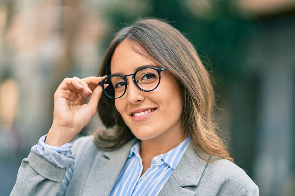 Junge hispanische Geschäftsfrau lächelt glücklich und berührt ihre Brille in der Stadt. - Foto, Bild