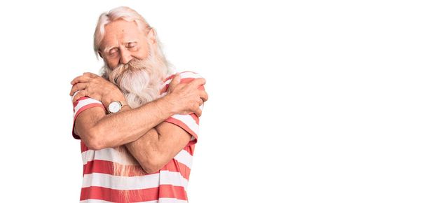 Vieil homme âgé aux cheveux gris et à la barbe longue portant un t-shirt rayé se serrant dans ses bras heureux et positifs, souriant confiant. amour de soi et soins de soi  - Photo, image