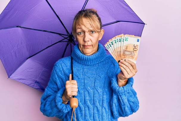 Middle age blonde woman holding umbrella and 50 euros banknotes clueless and confused expression. doubt concept.  - Photo, Image
