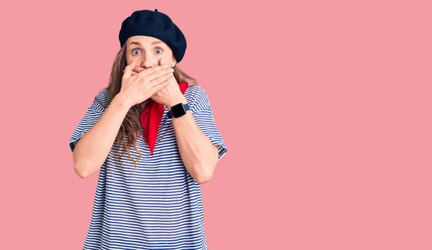 Young beautiful blonde woman wearing french beret and striped t-shirt shocked covering mouth with hands for mistake. secret concept.  - Photo, Image