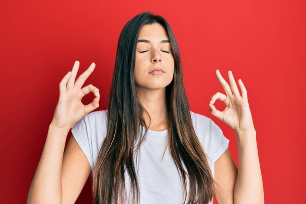 Jeune femme brune portant un t-shirt blanc décontracté sur fond rouge se détendre et sourire les yeux fermés en faisant un geste de méditation avec les doigts. concept de yoga.  - Photo, image