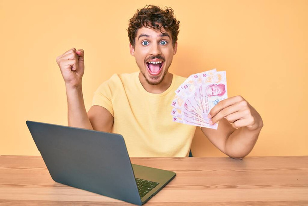 Young caucasian man with curly hair sitting on the table working with laptop and holding mexican pesos banknotes screaming proud, celebrating victory and success very excited with raised arms  - Photo, Image