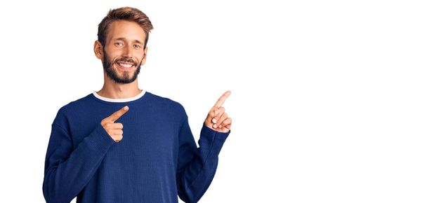 Hezký blonďatý muž s vousy na sobě ležérní svetr s úsměvem a při pohledu na kameru ukazuje dvěma rukama a prsty na stranu.  - Fotografie, Obrázek