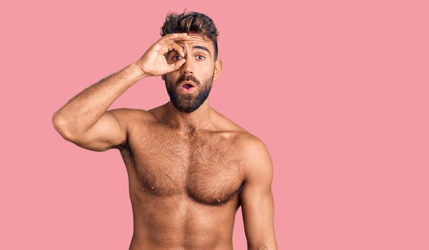 Young hispanic man wearing swimwear shirtless doing ok gesture shocked with surprised face, eye looking through fingers. unbelieving expression.  - Foto, Imagen