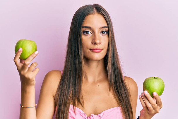 Jonge Spaanse vrouw met groene appels ontspannen met een serieuze uitdrukking op haar gezicht. eenvoudig en natuurlijk kijkend naar de camera.  - Foto, afbeelding